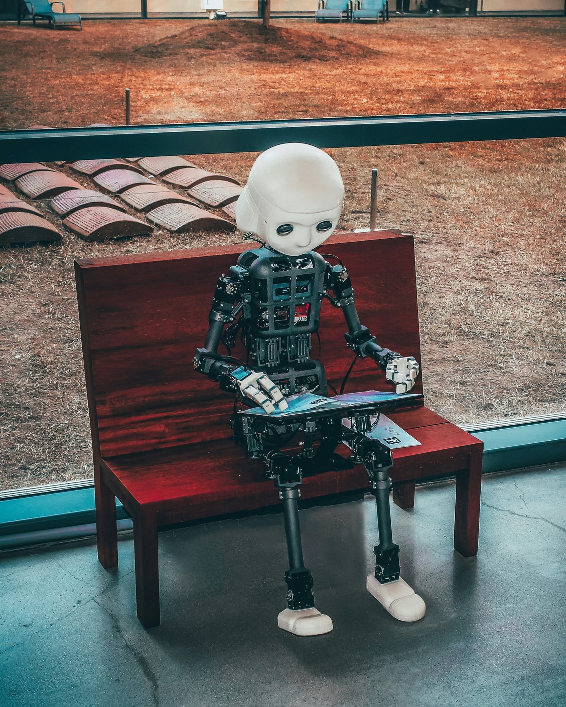 A small humanoid robot seated on a wooden bench holding a tablet computer